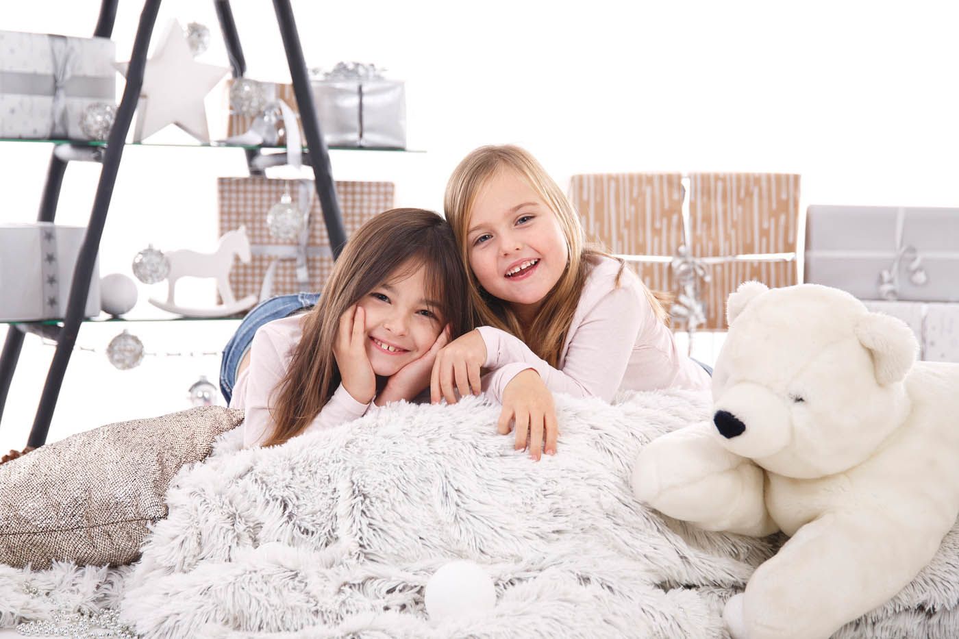 Zwei Mädchen lächeln und posieren auf einer flauschigen weißen Decke mit einem weißen Teddybären