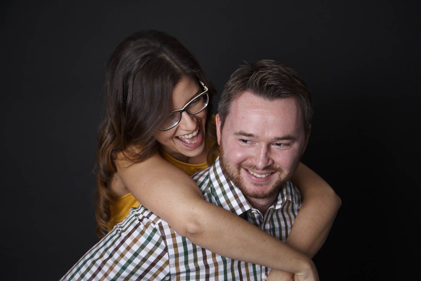 Eine lachende Frau umarmt einen Mann von hinten, beide lächeln vor schwarzem Hintergrund.