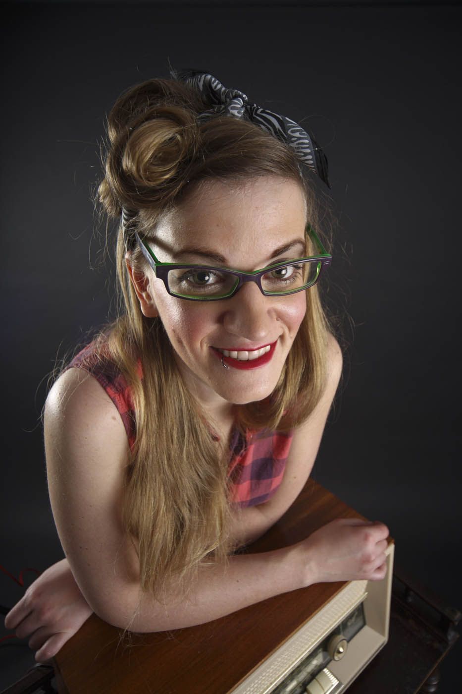 Eine Frau mit Brille lächelt und lehnt an einem Radio. Sie trägt ein kariertes Oberteil und hat ihr Haar mit einem Bandana hochgesteckt.