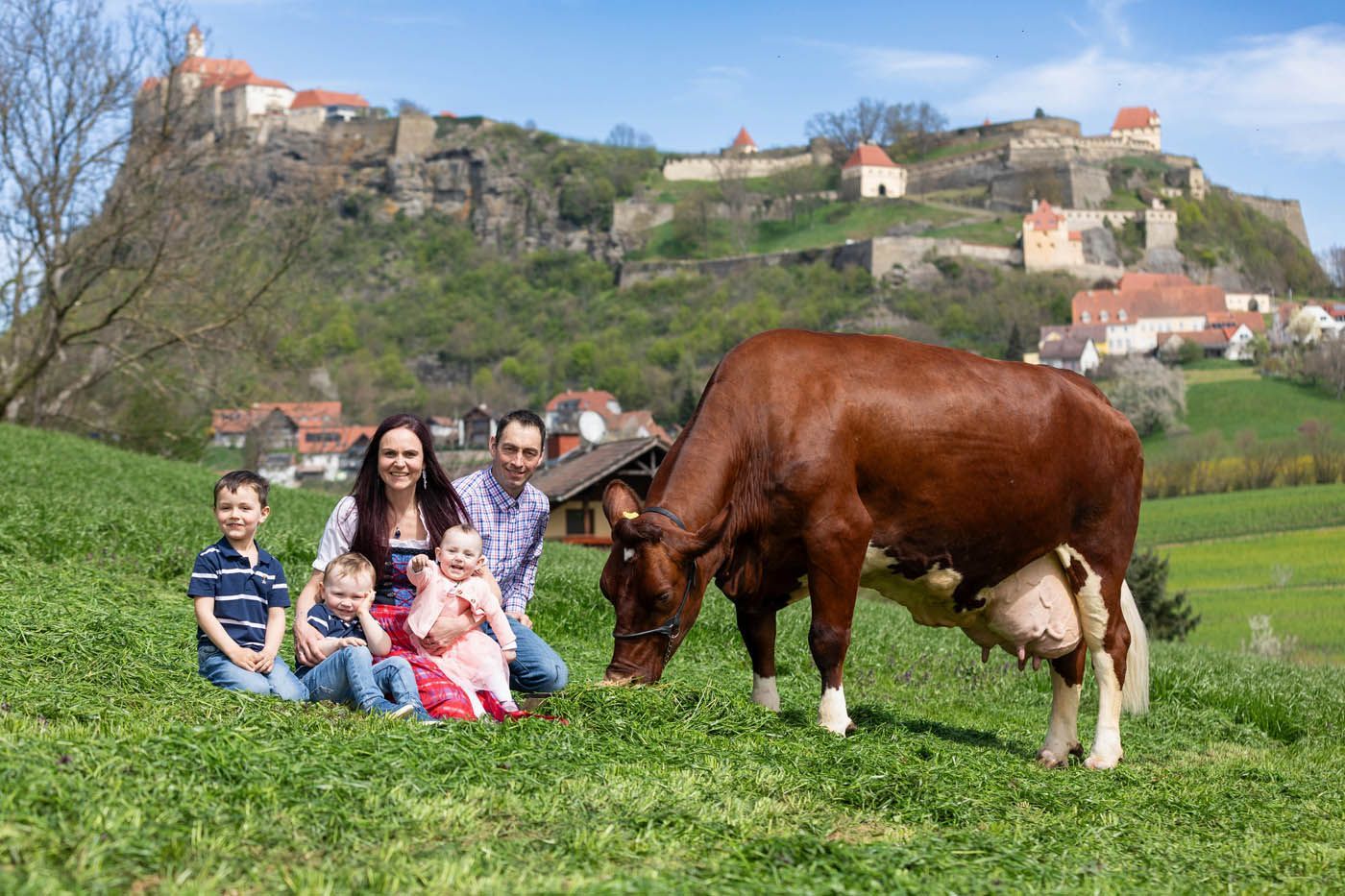 Eine Familie mit drei Kindern und einer Kuh grast vor einem Schloss auf einem Hügel.