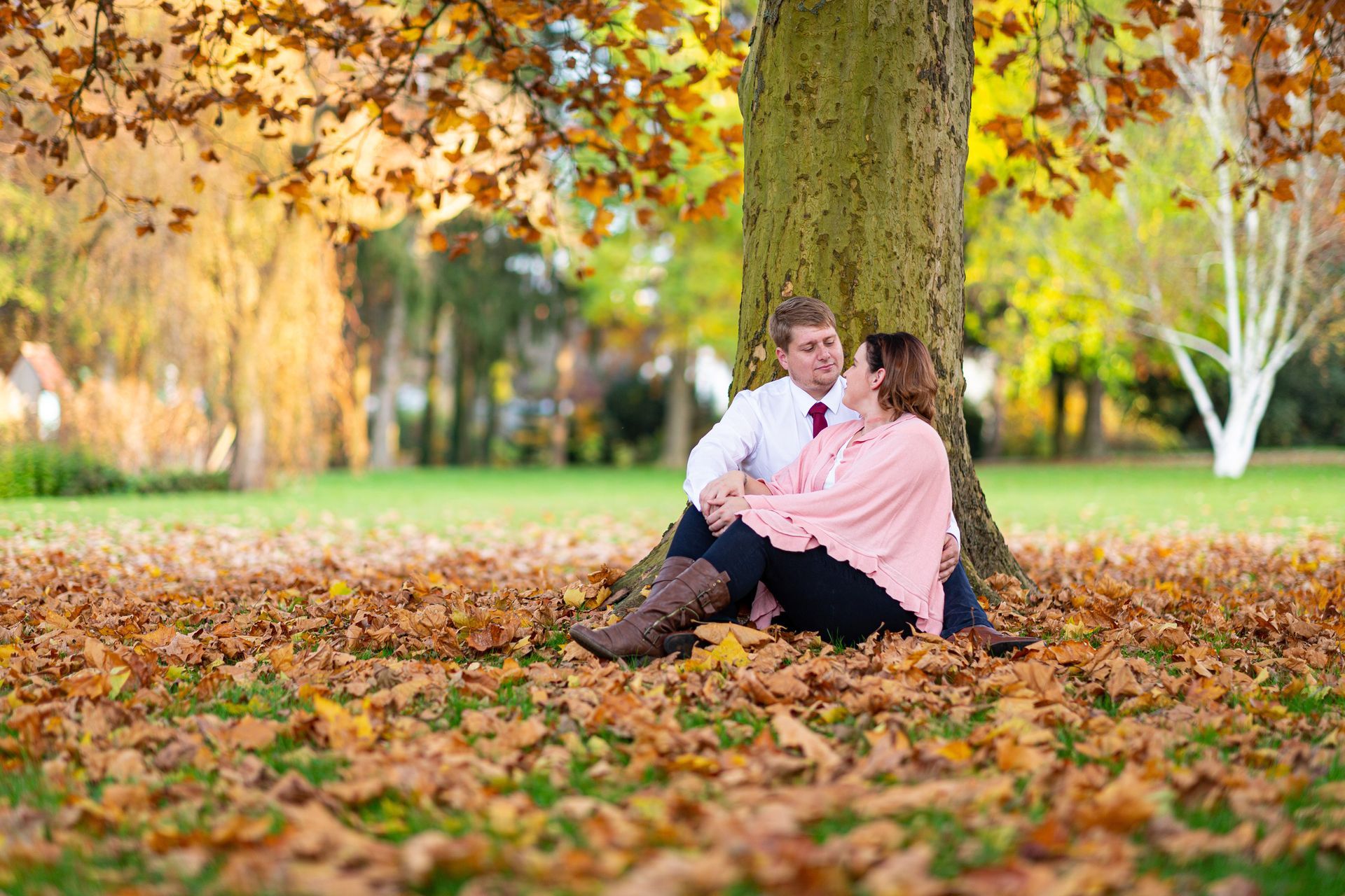 Ein Paar sitzt in einem Park unter einem Baum, umgeben von herabgefallenen Blättern.