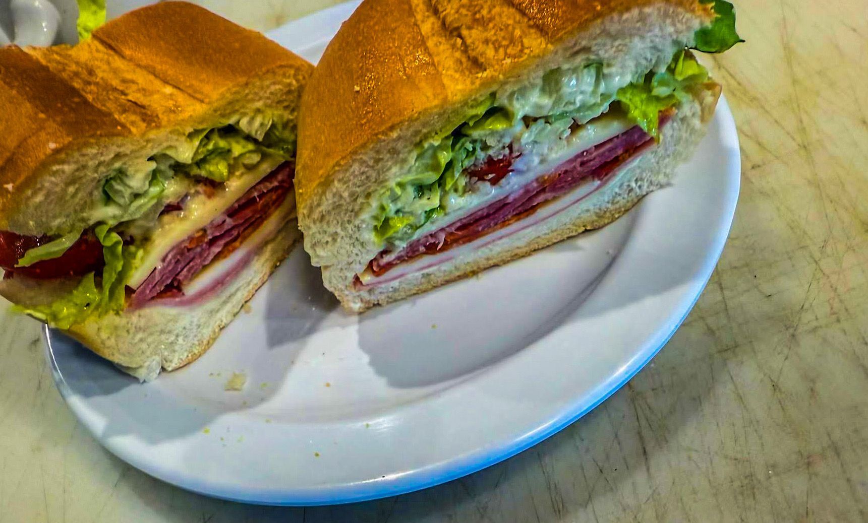 A large sandwich cut in half on a white plate; contains deli meats, cheese, lettuce, and tomatoes.