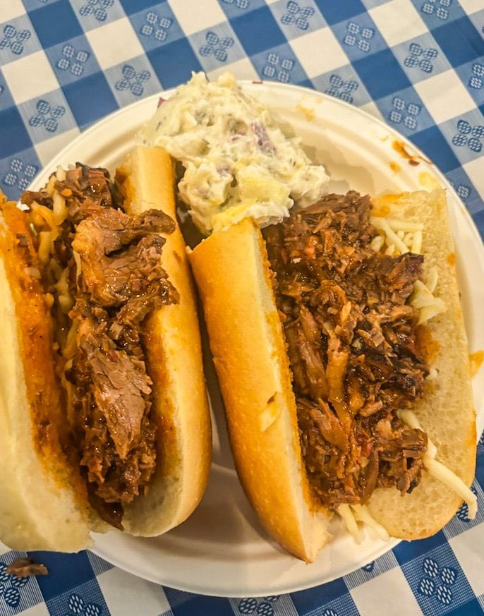 Two sandwiches with pulled meat and potato salad on a paper plate, blue checkered tablecloth.