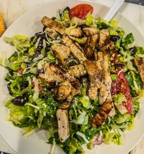 Grilled chicken salad with mixed greens, tomatoes, olives, and parmesan cheese on a white plate.