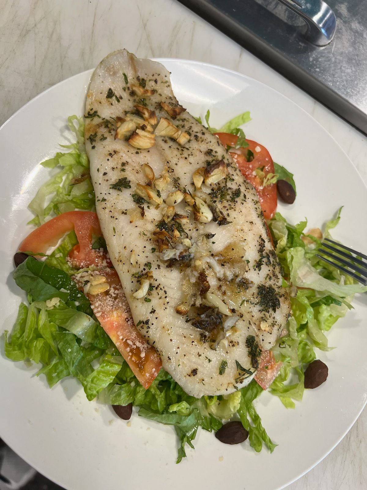Grilled fish fillet with herbs and pine nuts atop a salad of lettuce, tomatoes, and olives.