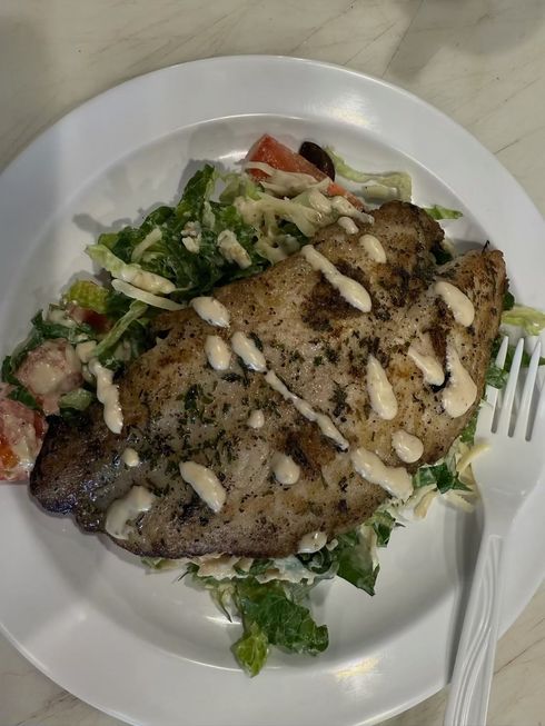 Grilled fish fillet drizzled with sauce over salad on a white plate, beside a plastic fork.