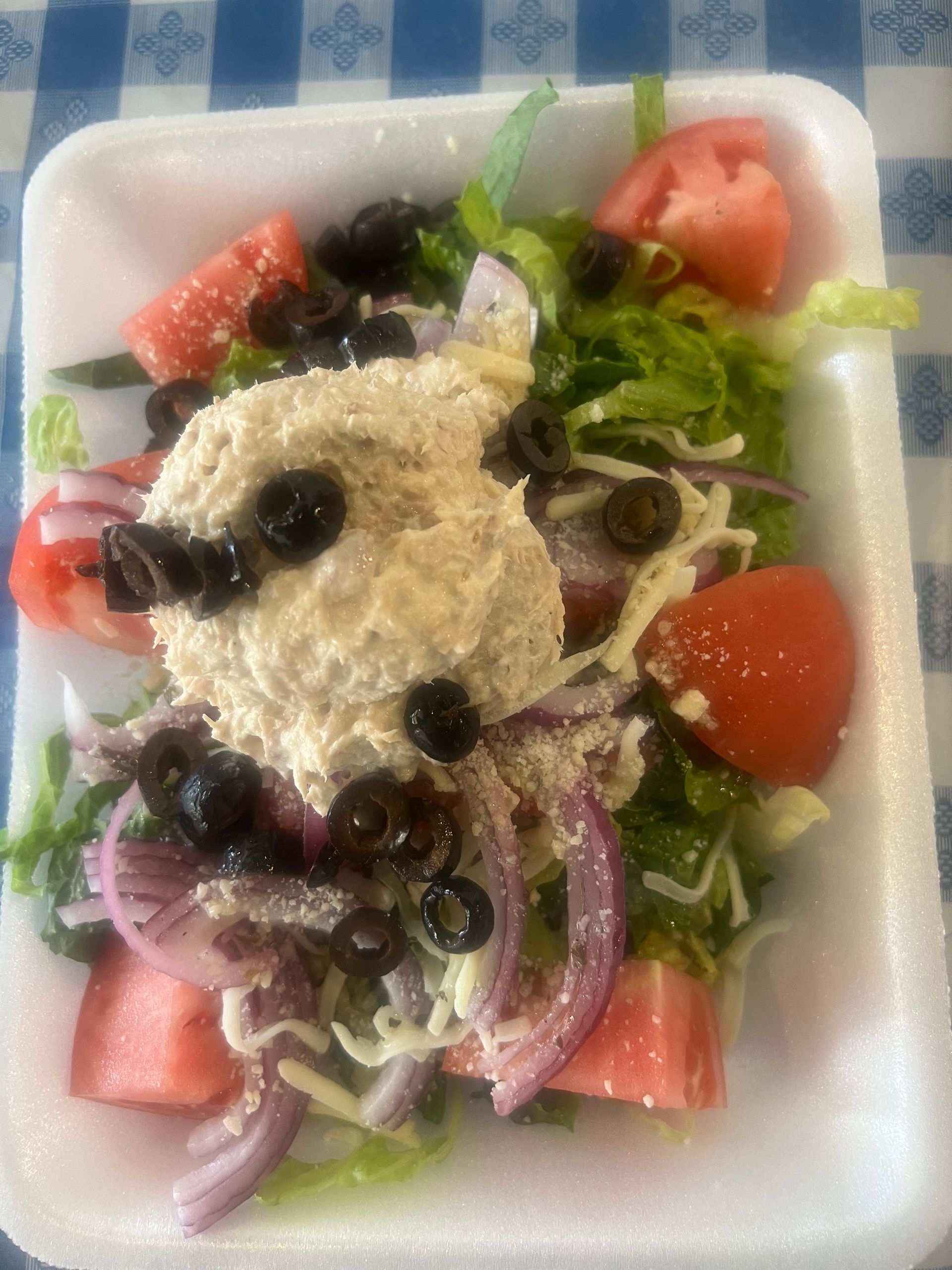 Salad in a white container, with tomatoes, olives, red onion, and a large dollop of creamy topping.