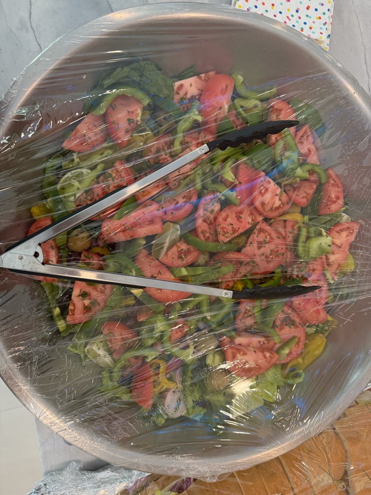 Large bowl of salad covered in plastic wrap with tongs resting on top.