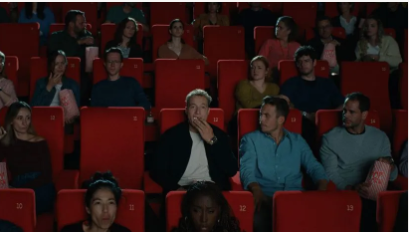 Publikum in einem Kino. Reihen roter Sitze, gefüllt mit unterschiedlichsten Menschen, die einen Film ansehen; manche sehen gelangweilt aus.