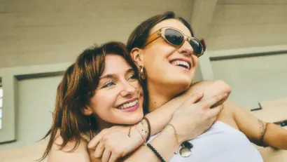 Zwei Frauen umarmen sich lächelnd im Freien. Eine trägt eine Sonnenbrille. Sonniger Tag, beiger Hintergrund.