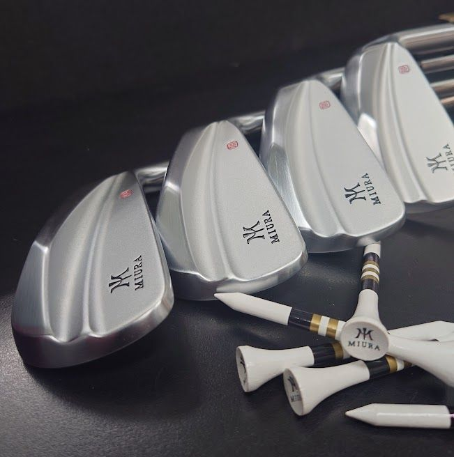 Twee mannen op een golfbaan, de ene met een club in de hand, de andere kijkend. Groen gras, golfkarretje, palmbomen.