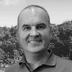 Man met kort haar en een lichte glimlach, gekleed in een poloshirt, met bomen op de achtergrond.