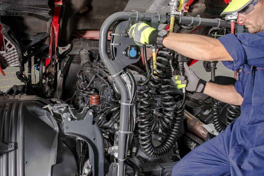 Mechanic in blue coveralls repairing a truck engine with hoses and wiring exposed