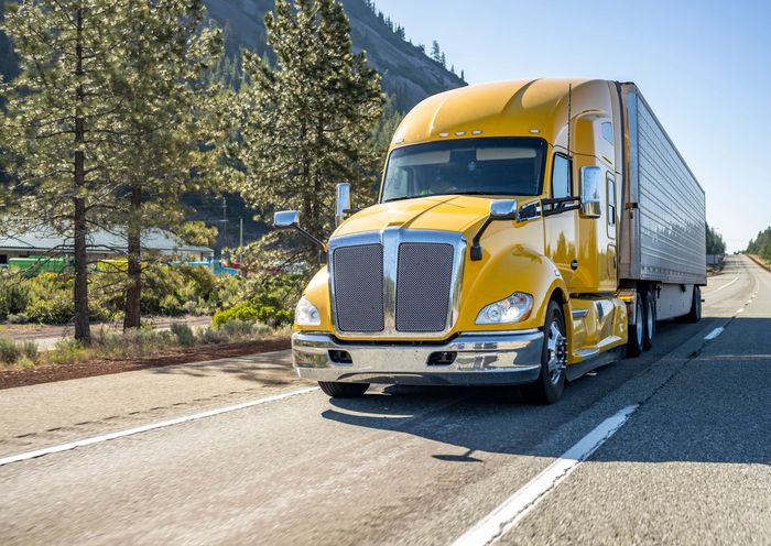 Yellow semi-truck driving on a sunny highway beside trees and mountains