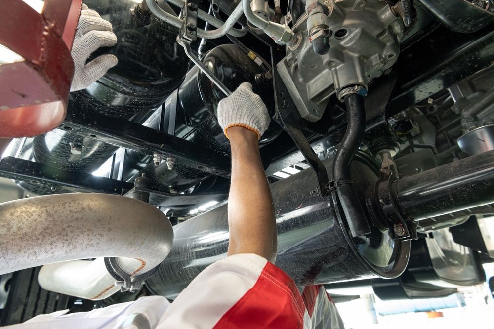 Mechanic inspecting machinery under a vehicle, reaching up with a gloved hand in an auto repair bay