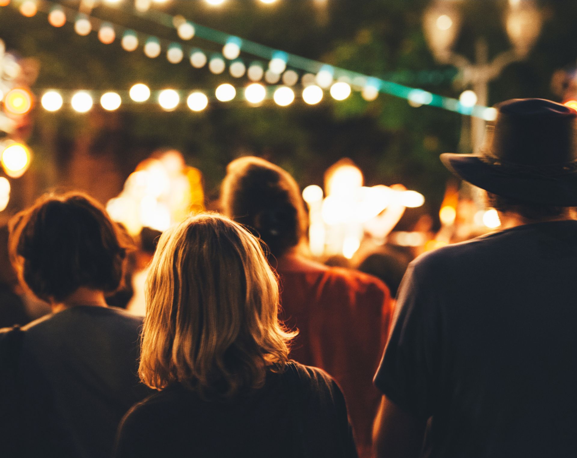 People at an outdoor event at night, looking toward stage; string lights and blurred figures in the background.