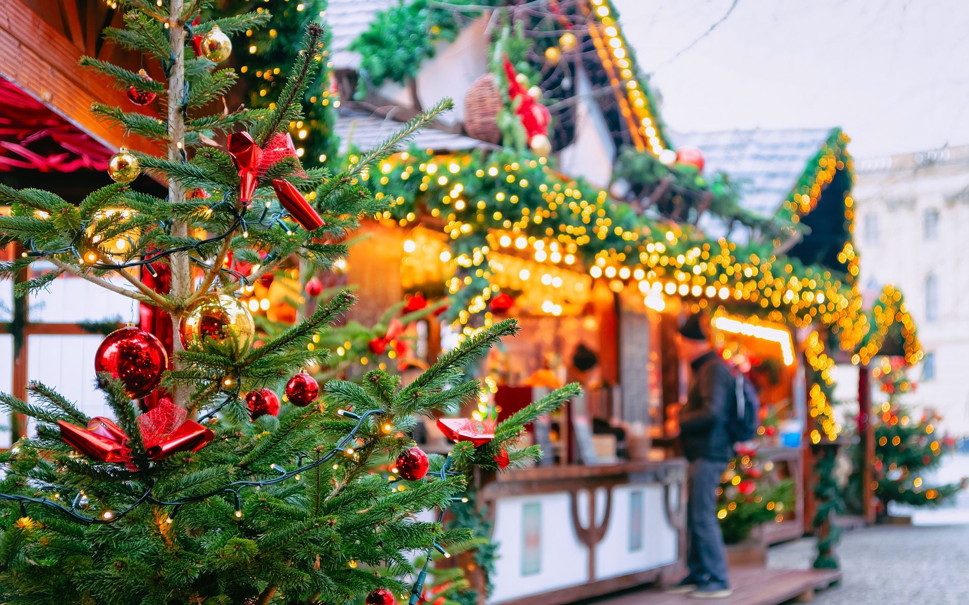 Christmas market scene: decorated tree in foreground, stalls with lights, person looking at a stall.