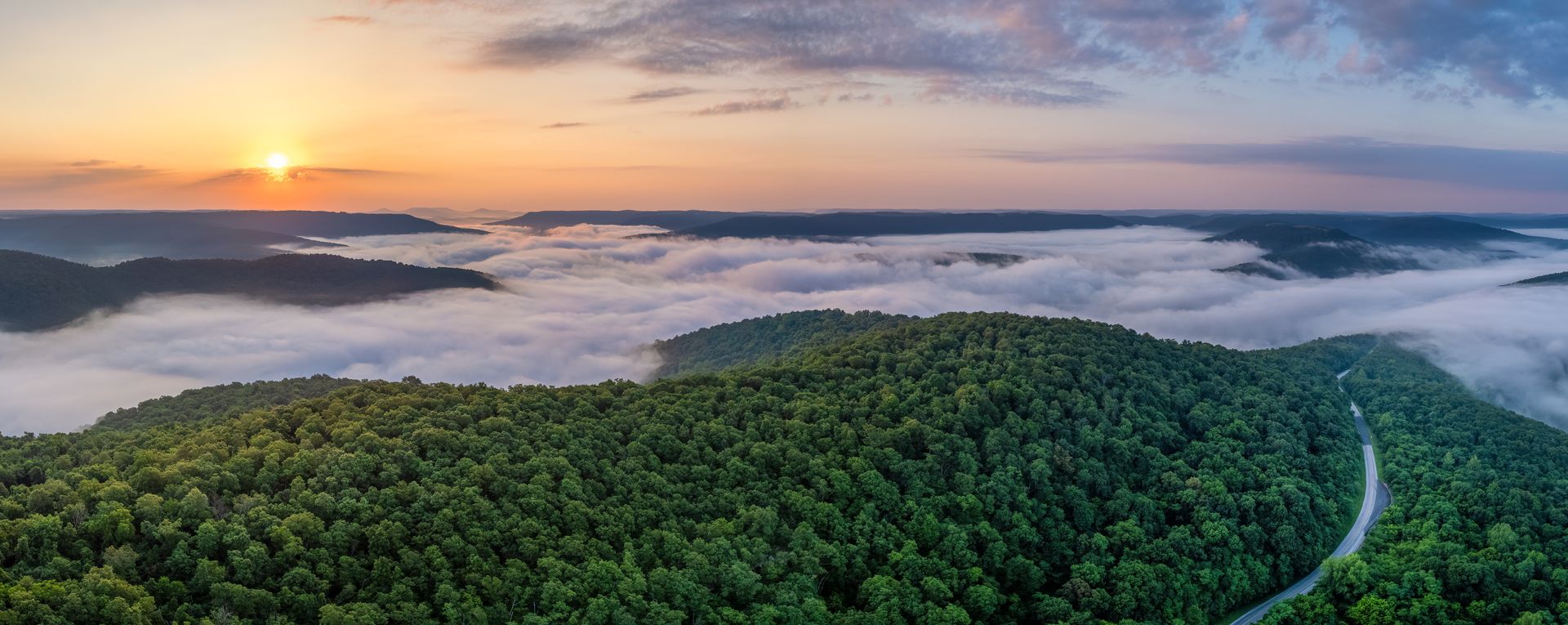 Aerial view of a mountain range covered in green trees, with fog filling the valleys, at sunset.