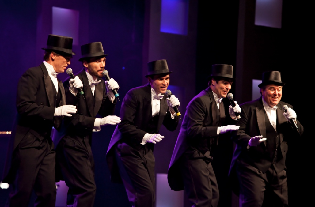 Four men in tuxedos and top hats are singing into microphones on a stage