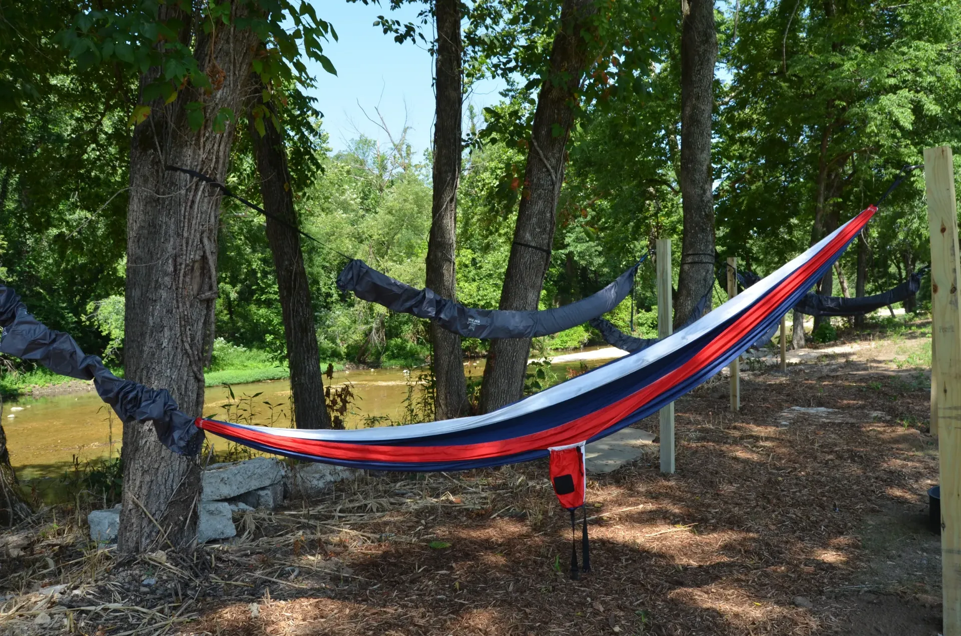 A red , white and blue hammock is hanging between two trees in the woods.