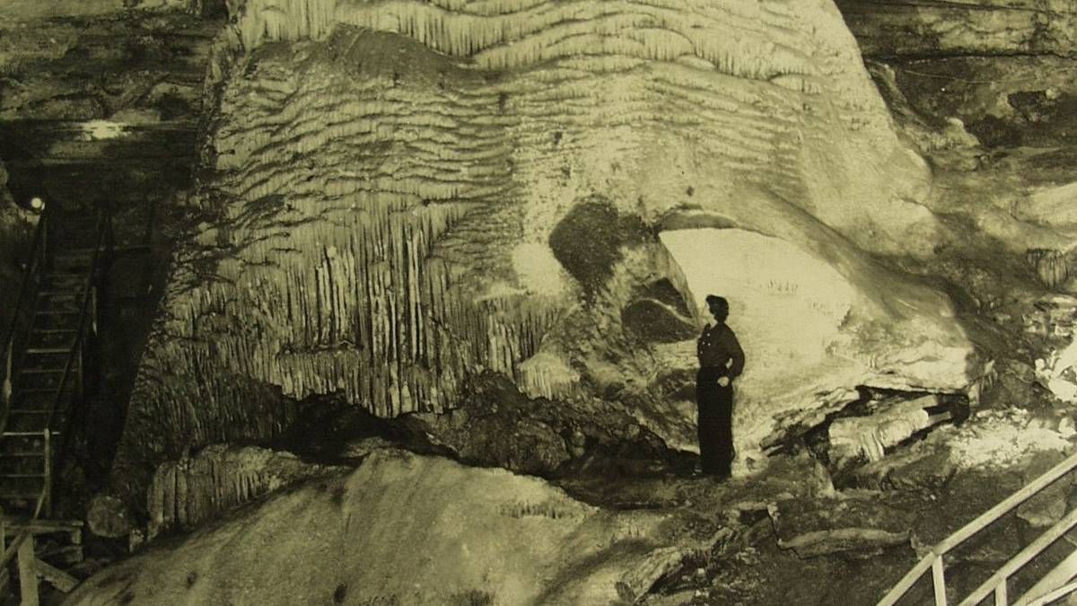 Person standing in a large cave, looking up at layered rock formations and stalactites.