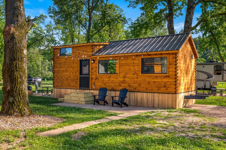 Small, wood-paneled cabin with a metal roof. Two chairs sit on a small patio in a grassy area.