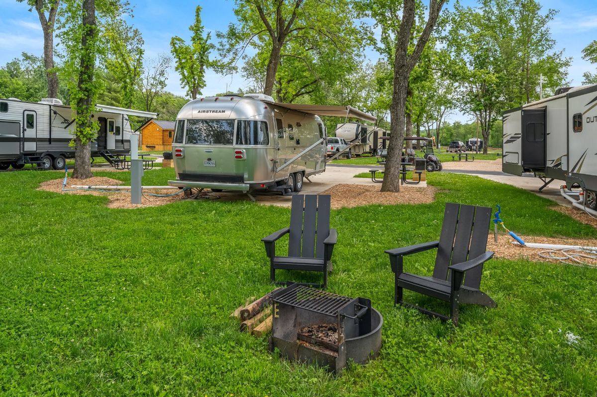 A rv park with a fire pit and chairs in the grass.