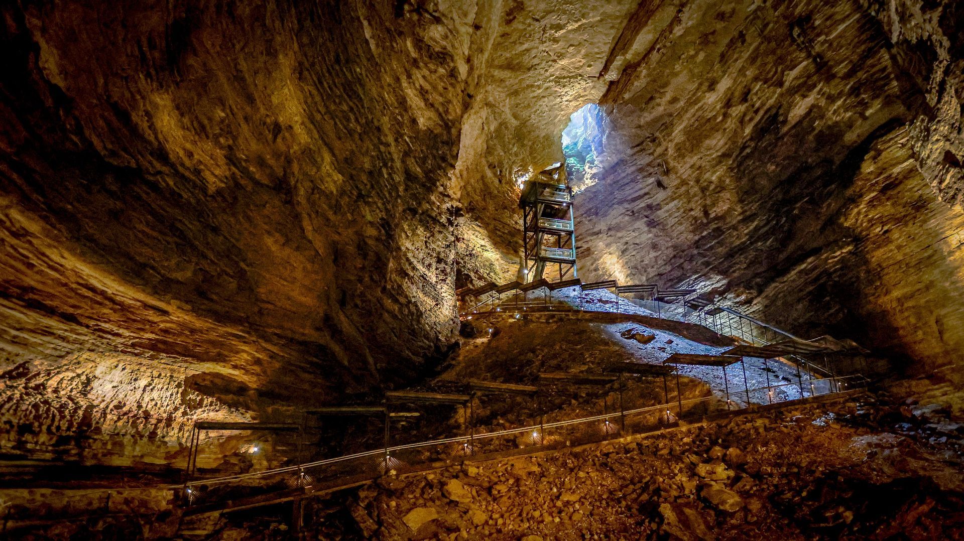 Inside a cave, lit path winds past rock formations towards an opening.