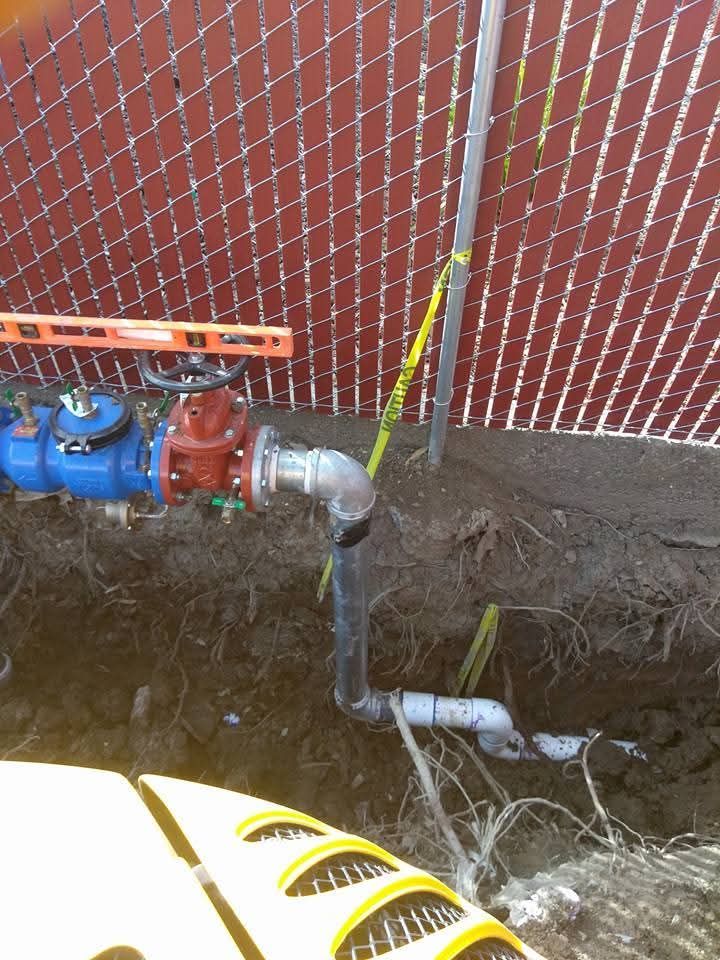 Pipes and valve assembly, partly buried, next to a chain-link fence, with water flowing from a joint.