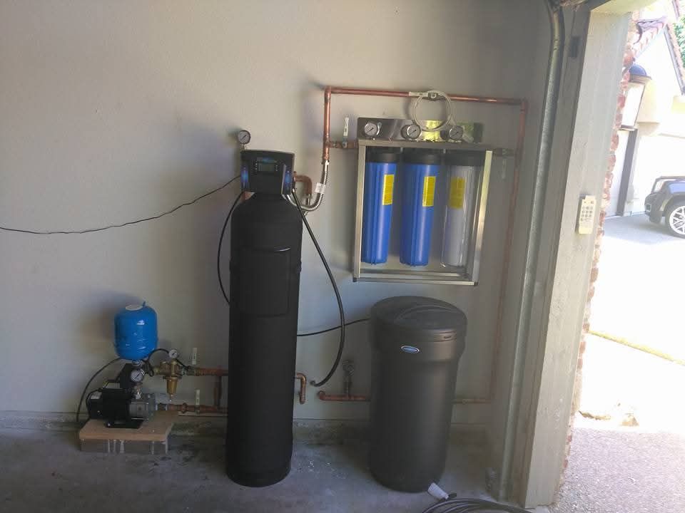 Water filtration system in a garage: black tanks, blue and gray filters, copper pipes, pump, blue pressure tank.