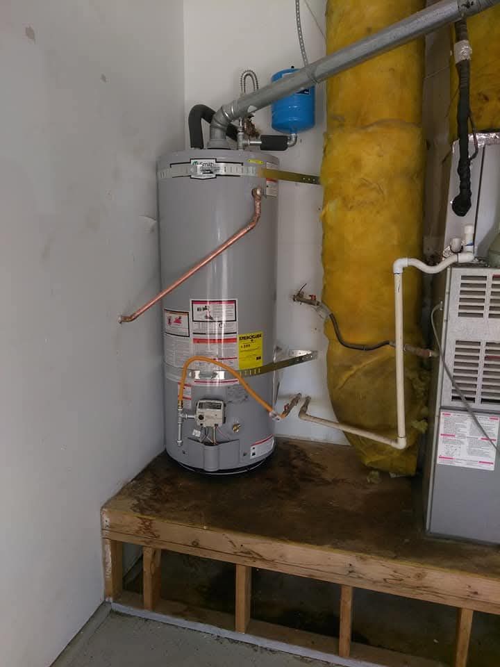 Water heater and associated pipes in a garage, mounted on a wooden frame, with a furnace and insulation nearby.