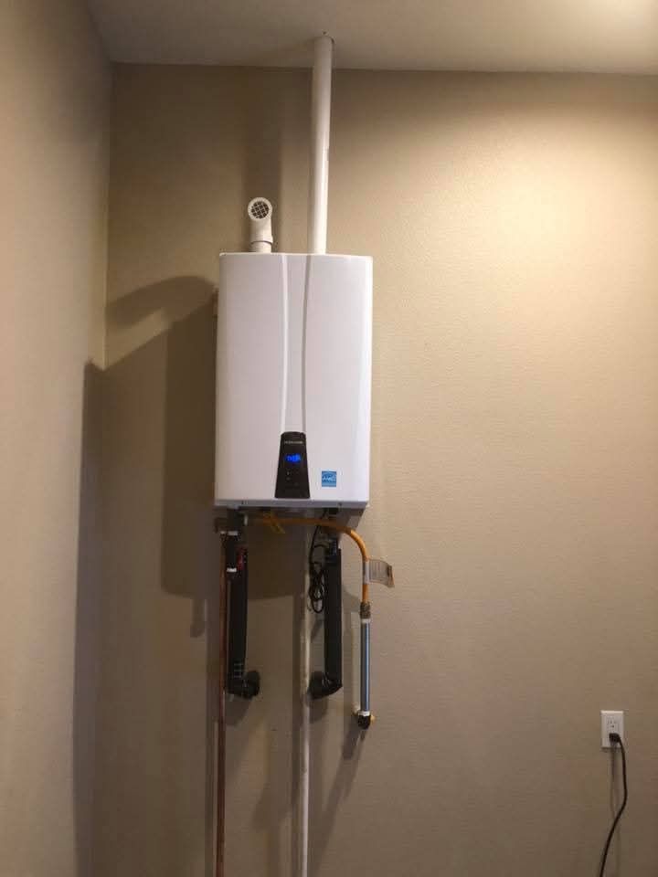 White tankless water heater mounted on a beige wall with venting pipes and plumbing visible.