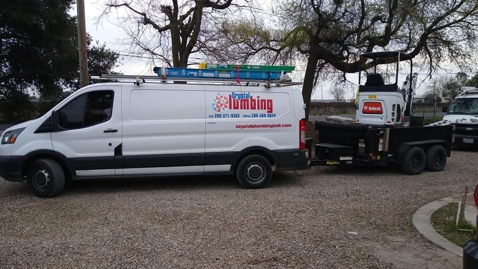 White plumbing van with logo, pulling a trailer with a small excavator on gravel driveway.