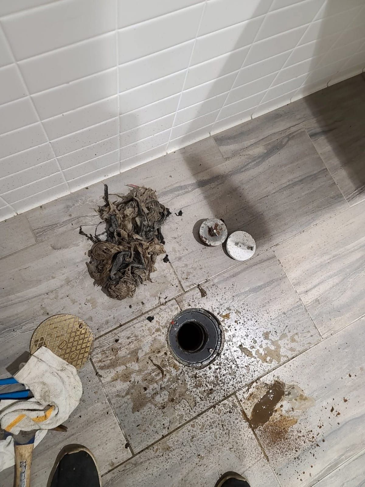 Shower drain with removed hair and debris on tiled floor, along with drain parts and cleaning tools.