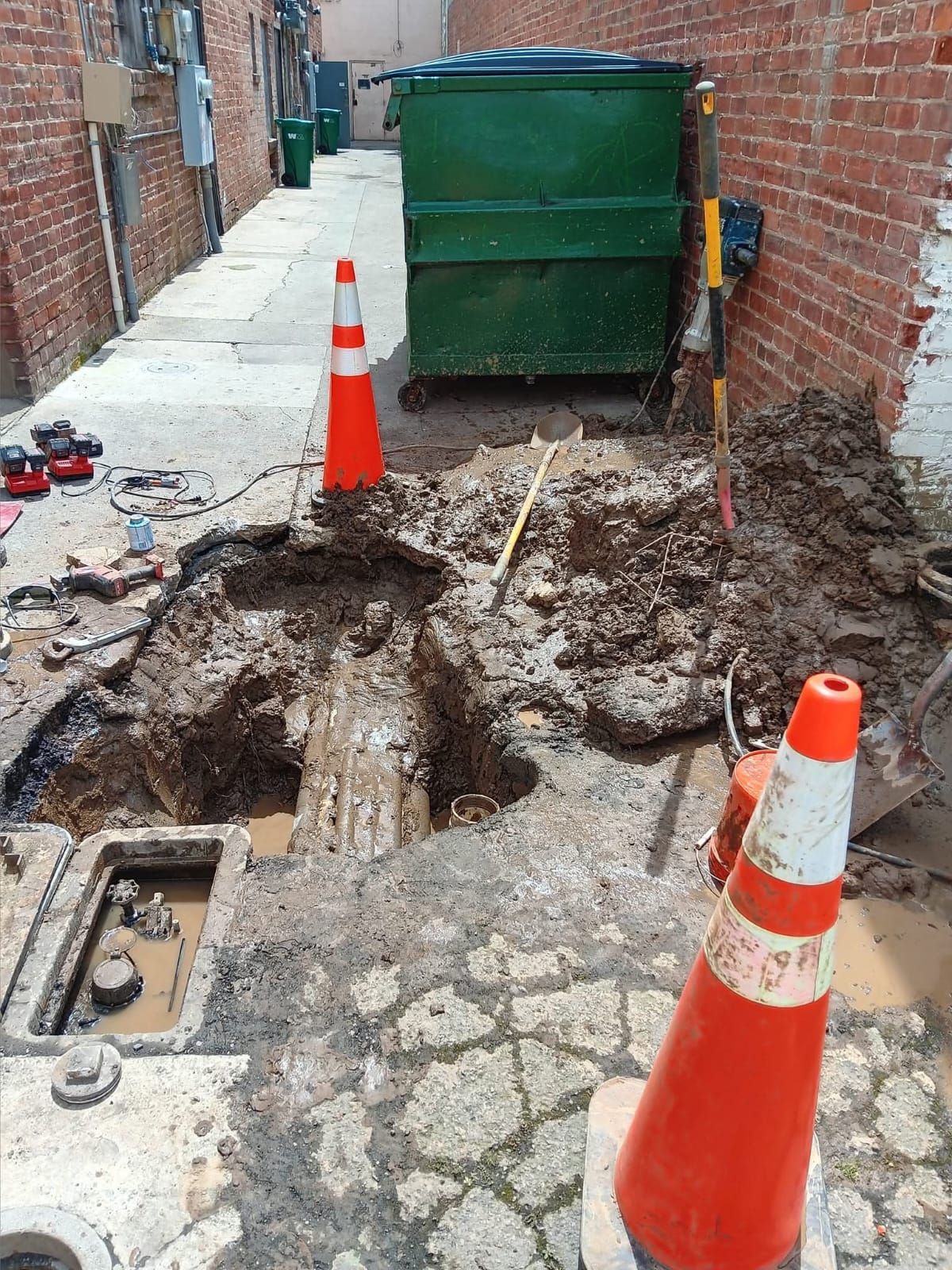 Construction site in an alley: open hole, orange cones, green dumpster, and brick walls.