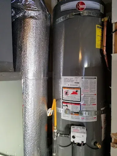 A silver Rheem water heater with a foil-wrapped duct next to it in a utility room.