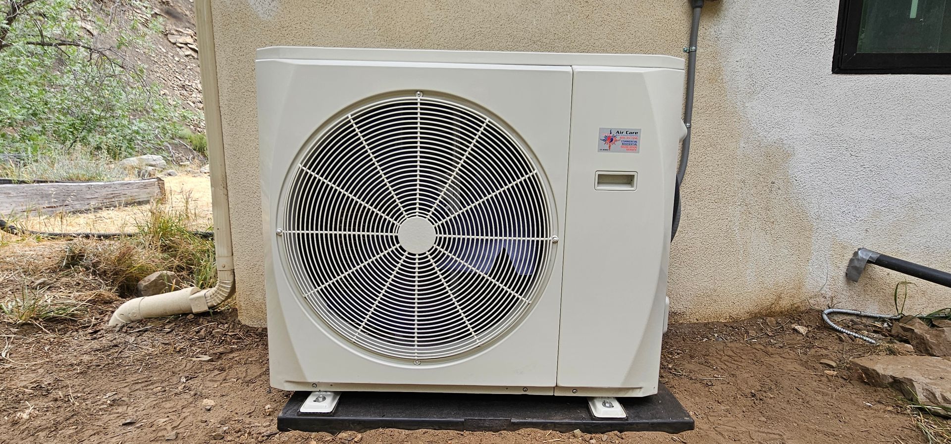 Outdoor air conditioner unit beside a beige wall on gravel, with a large circular fan grill.