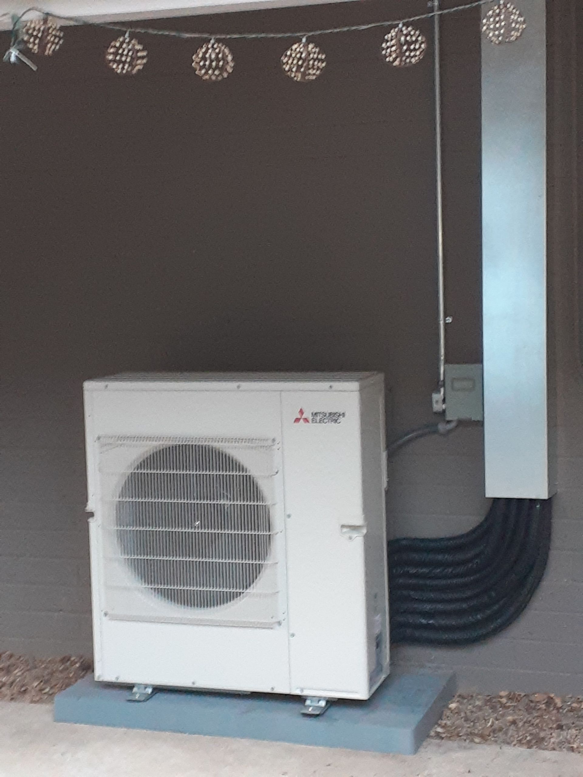 Outdoor air conditioner unit beside a wall, with black hoses and a gray metal panel overhead.