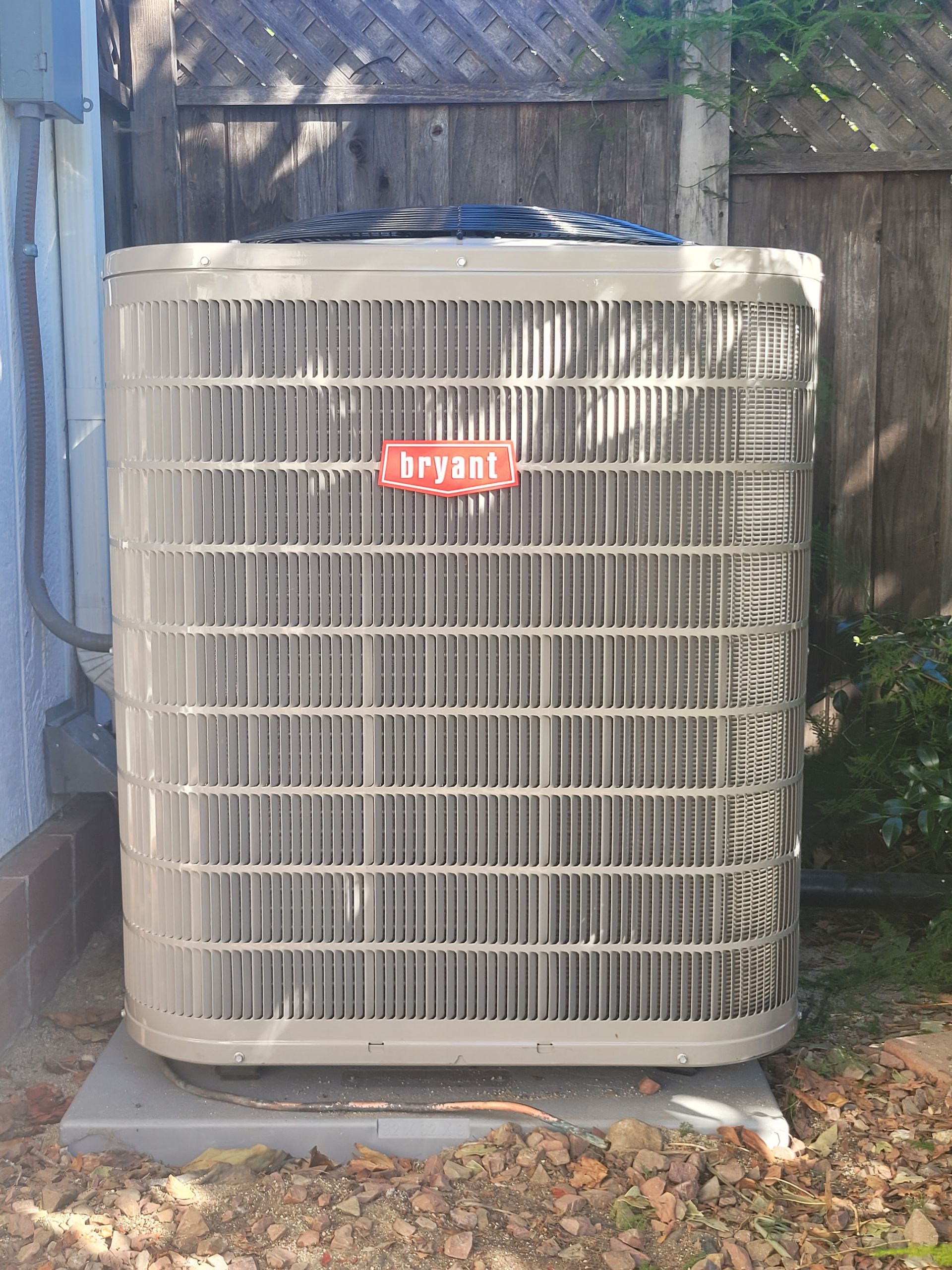 Large outdoor air conditioner unit against a fence, with a red logo and plants nearby.