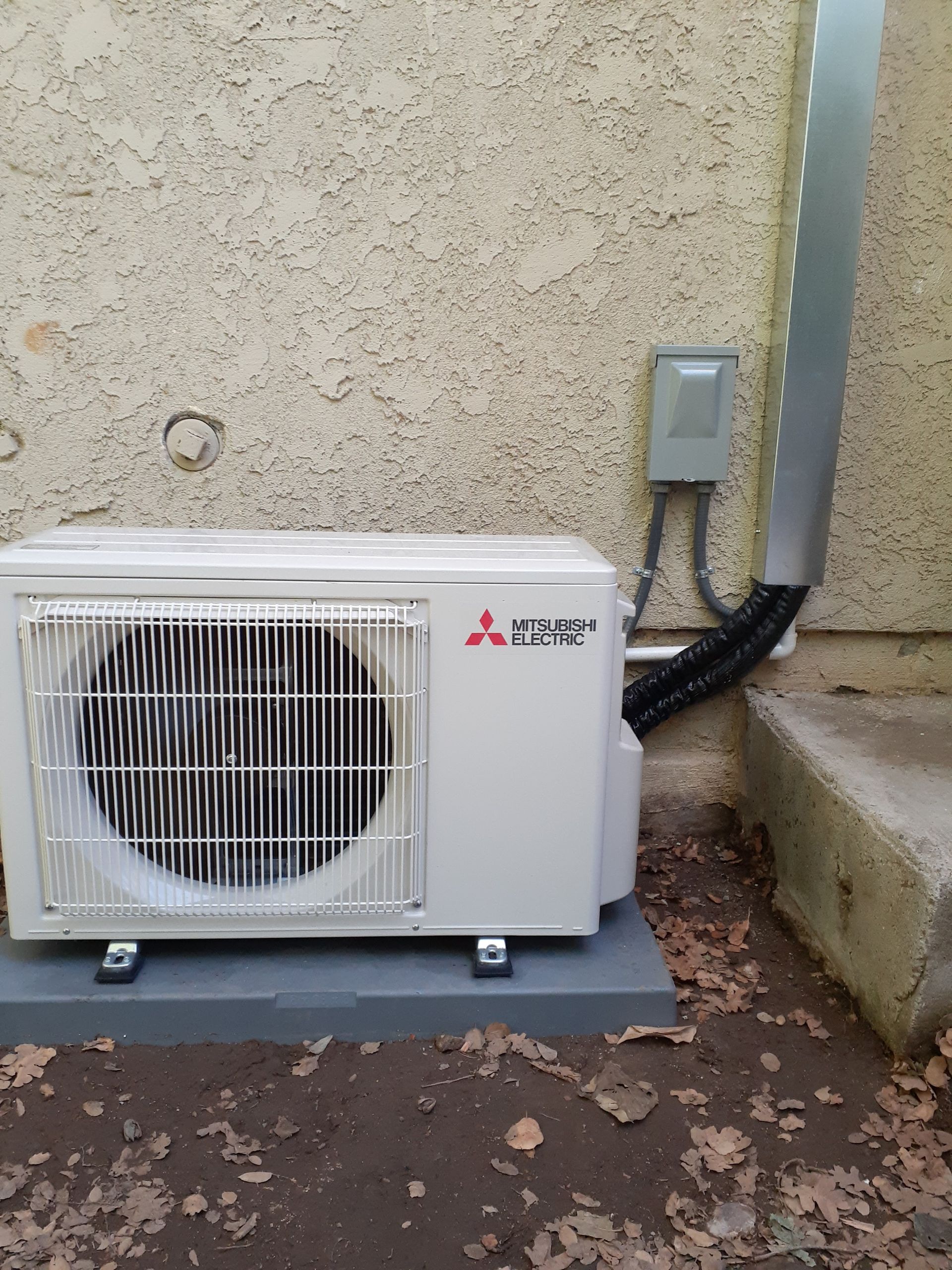 Outdoor Mitsubishi Electric heat pump unit beside a stucco wall and step, with conduit and control box.