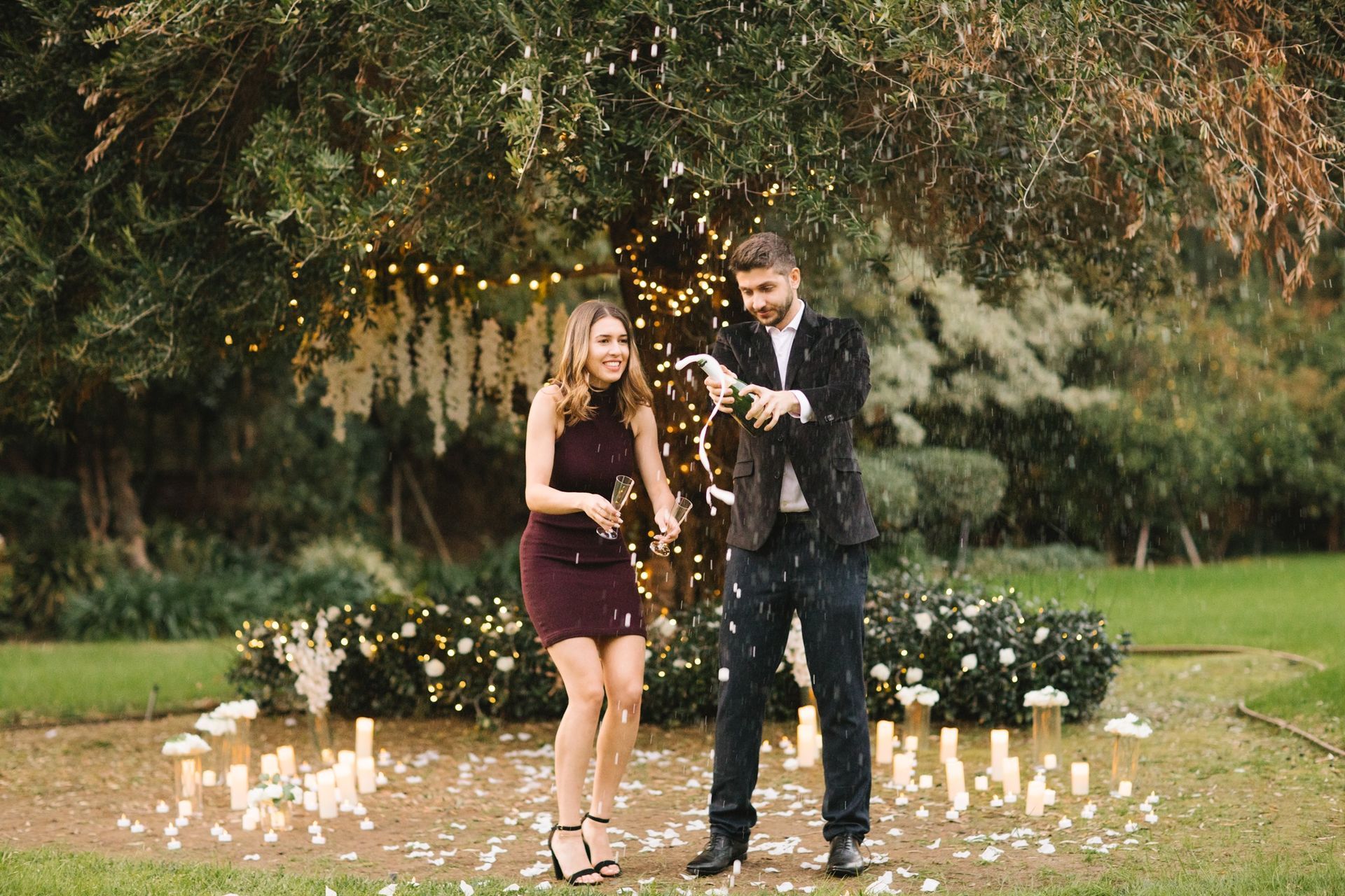A newly engaged couple laughing and popping a bottle of champagne, with bubbly spraying in the air and a joyful, celebratory atmosphere.