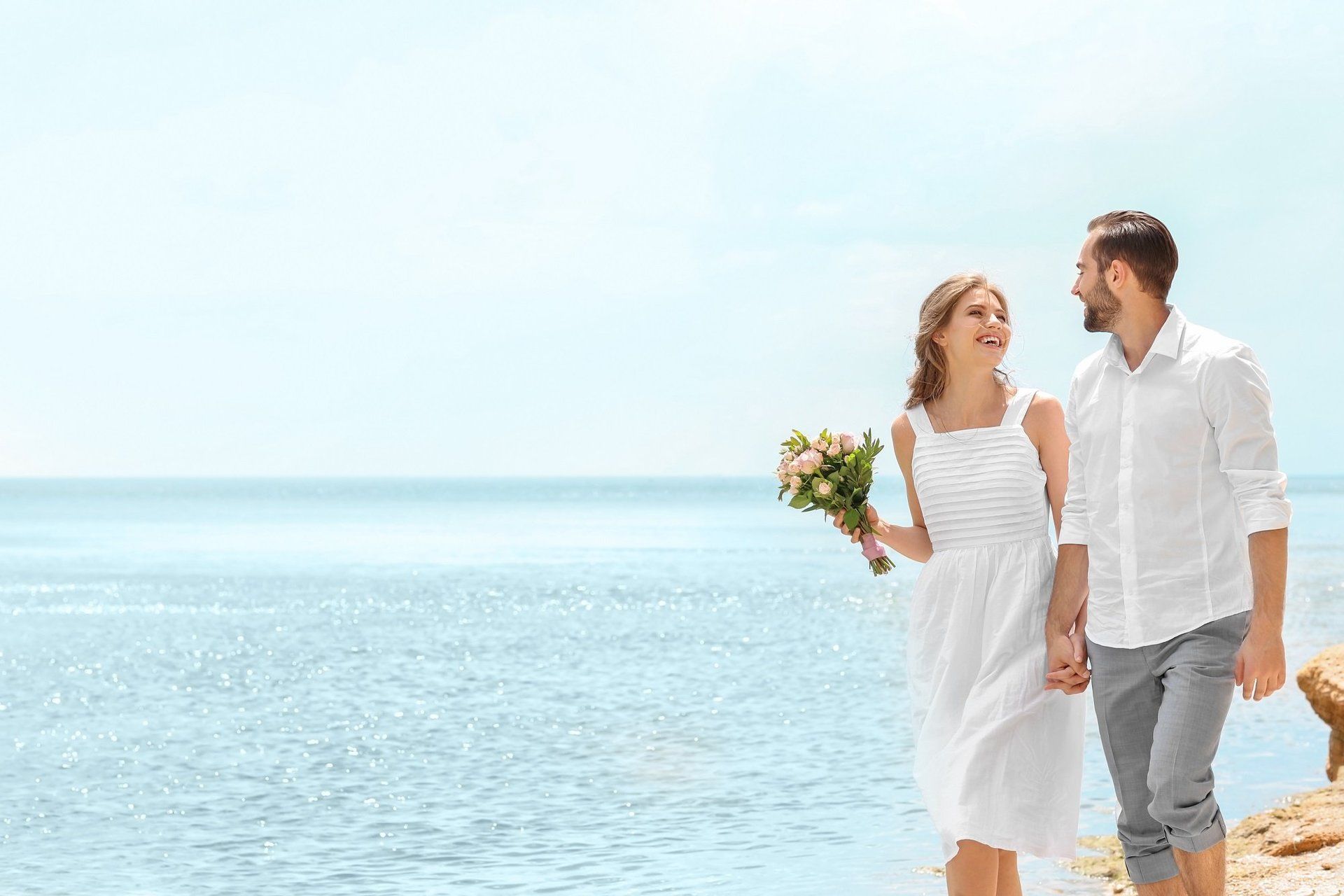 A bride and groom are walking on the beach holding hands.