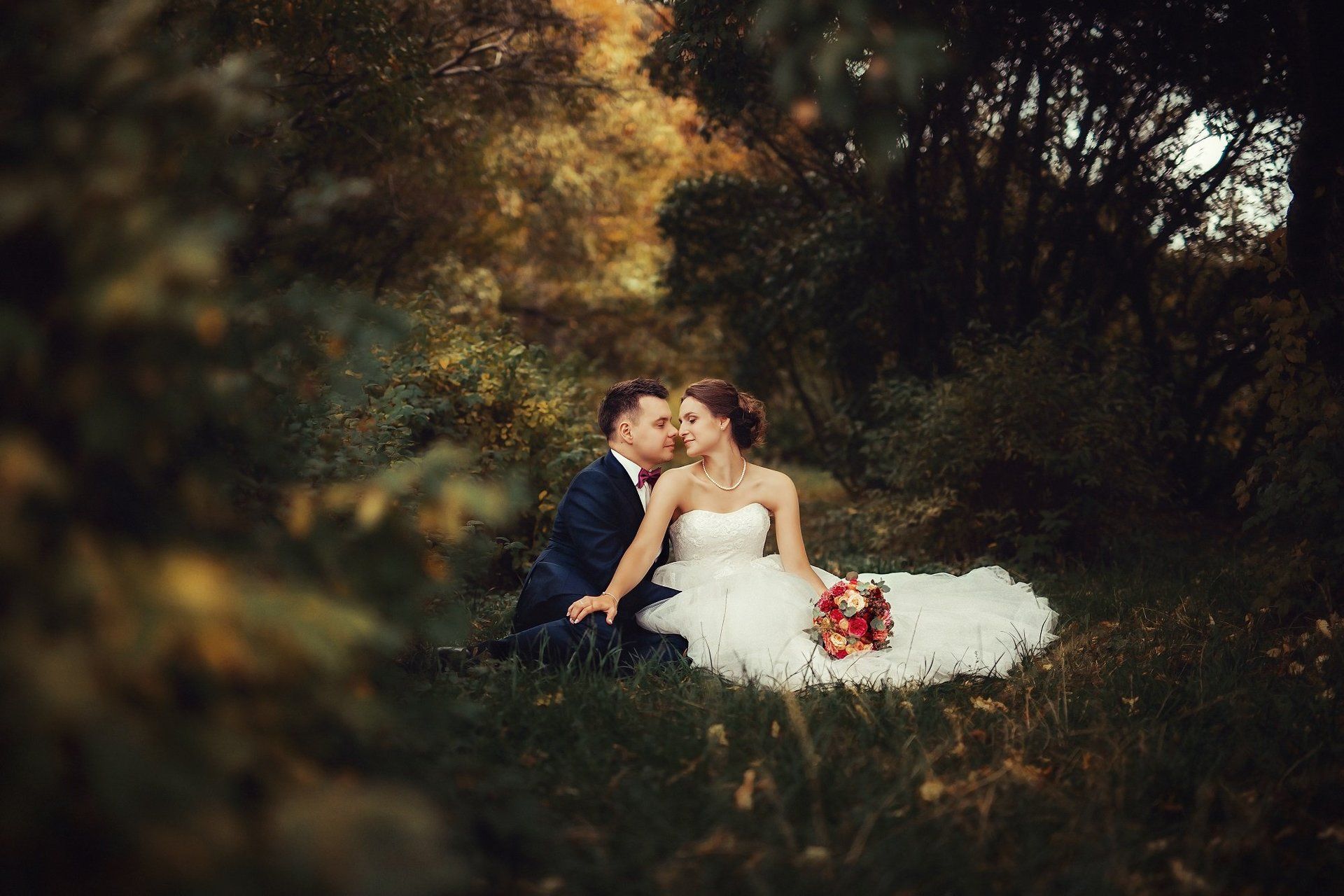 A bride and groom are sitting in the grass and kissing.