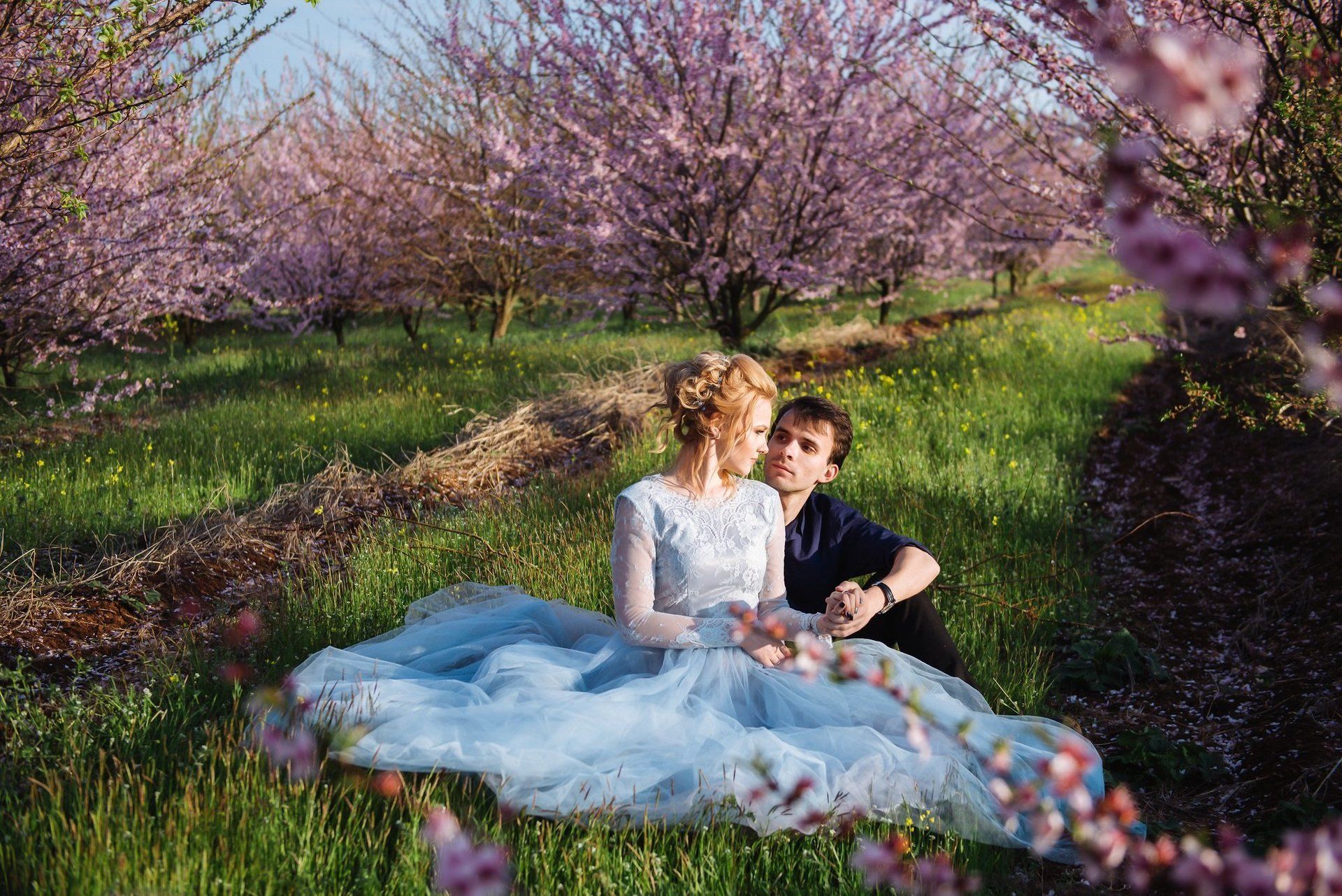 A man and a woman are sitting in a field of flowers . the woman is wearing a blue dress.