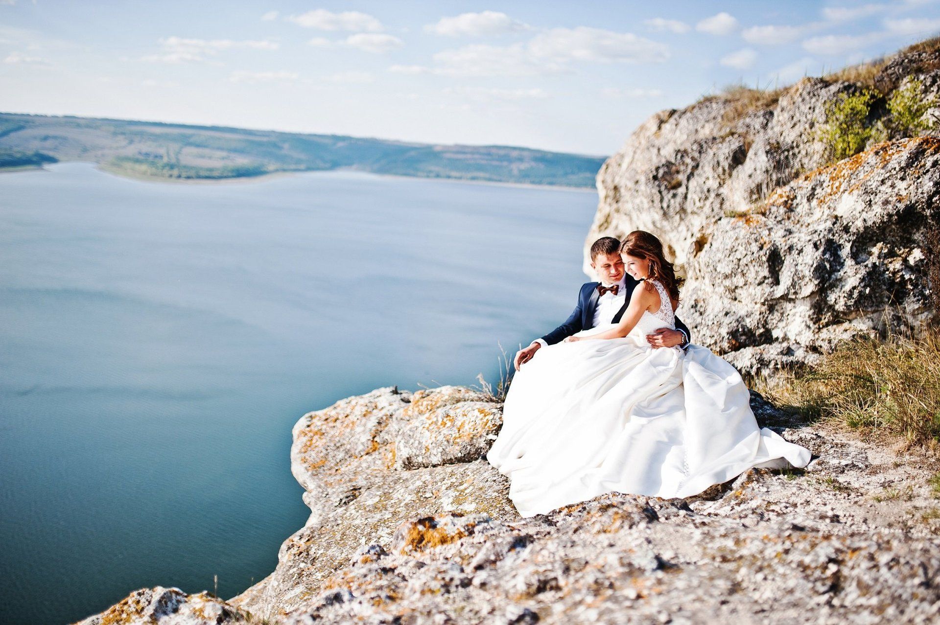 A bride and groom are sitting on the edge of a cliff overlooking a lake.