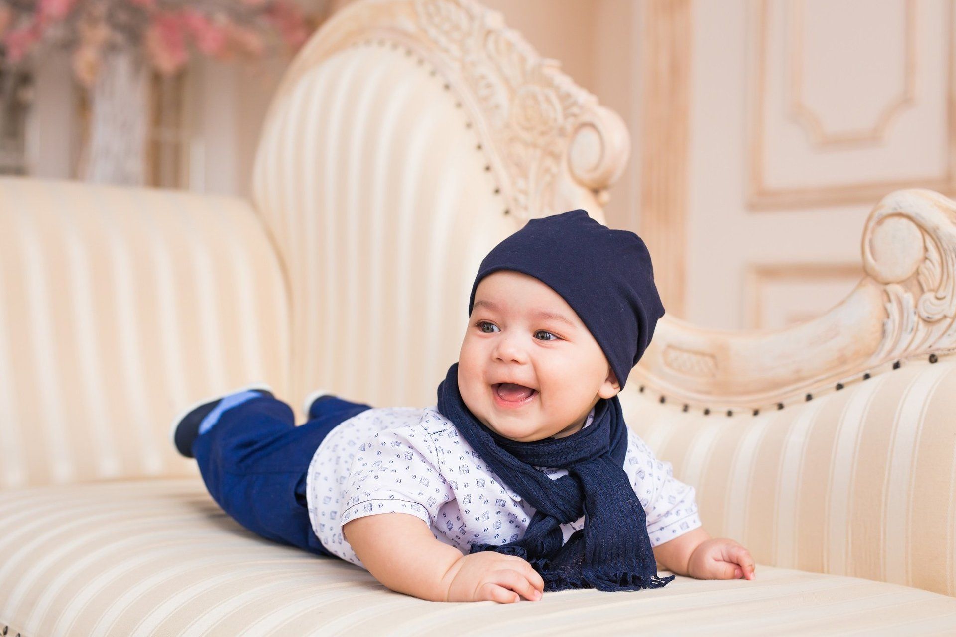 A baby is laying on his stomach on a couch wearing a hat and scarf.