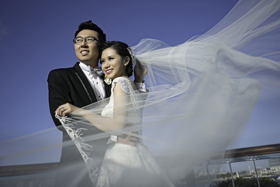 A bride and groom are posing for a picture with their veil blowing in the wind.