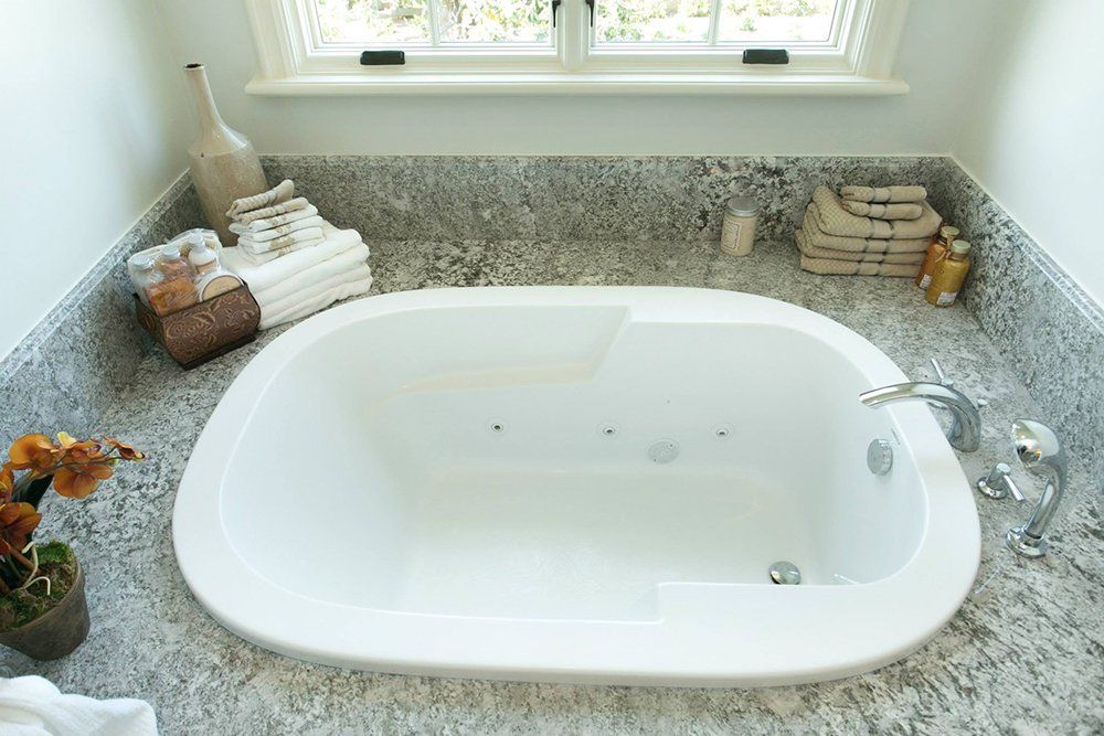 A bathtub is sitting on a granite counter in a bathroom next to a window.