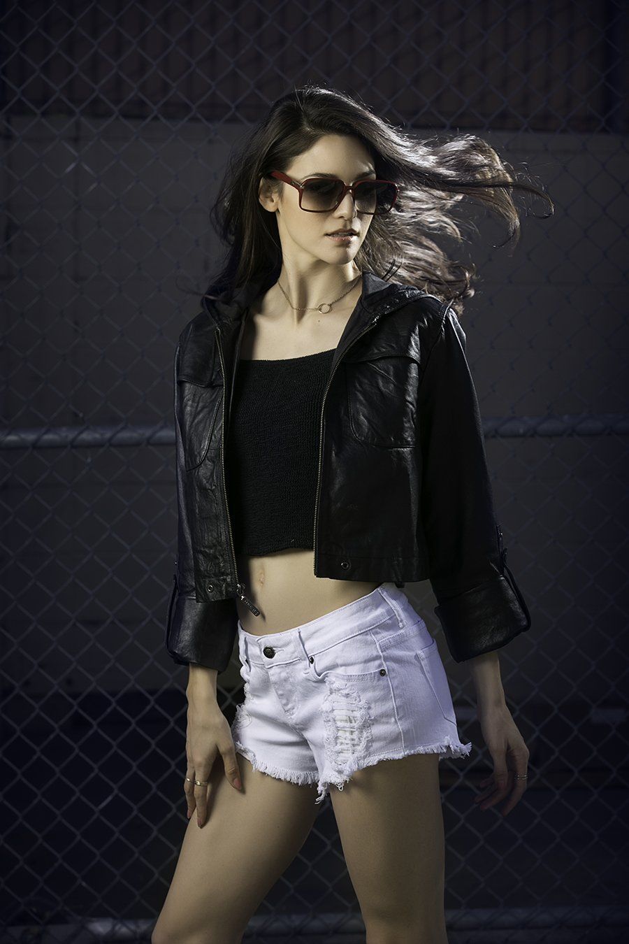 A woman wearing sunglasses and a black jacket is standing in front of a chain link fence.