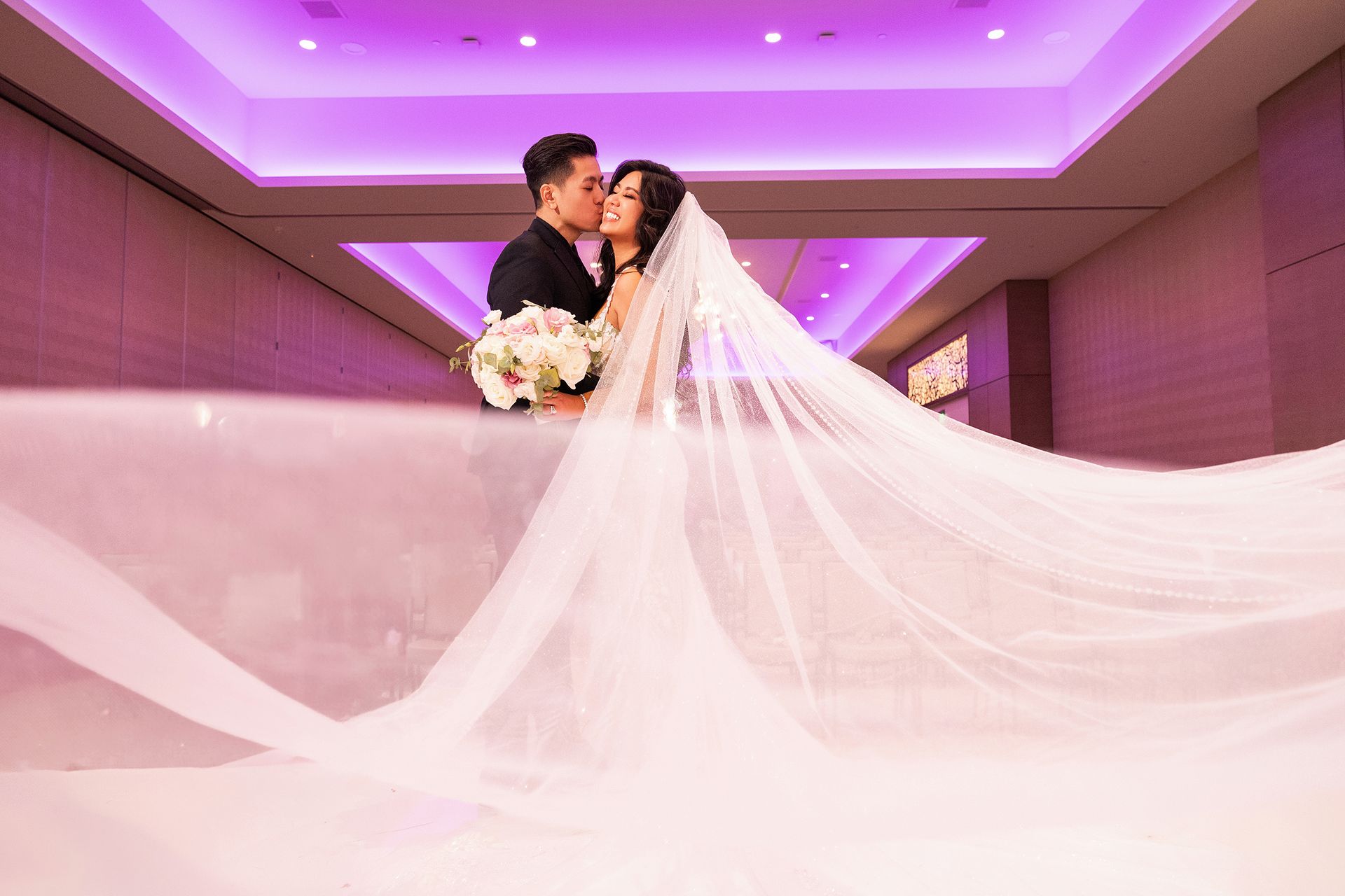 A bride and groom are kissing in a room with purple lights . the bride is wearing a long veil.